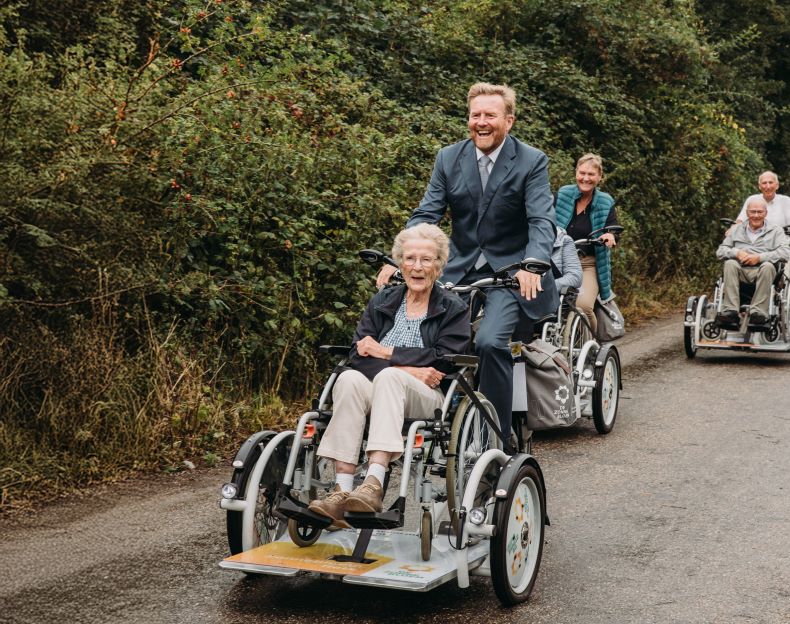 Koning Willem-Alexander op de Zonnebloemfiets met voorop een deelnemer. Achter hen nog twee duo's op de Zonnebloemfiets.